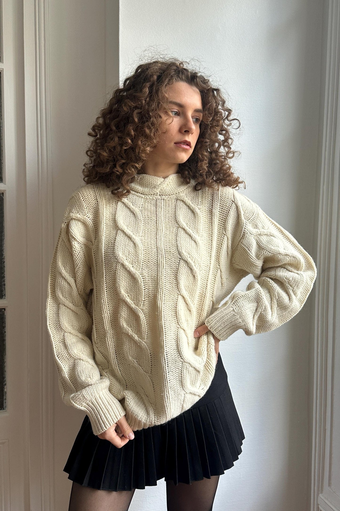 A young woman with curly hair stands in front of a white wall, wearing a cream-colored sweater and a black pleated skirt.