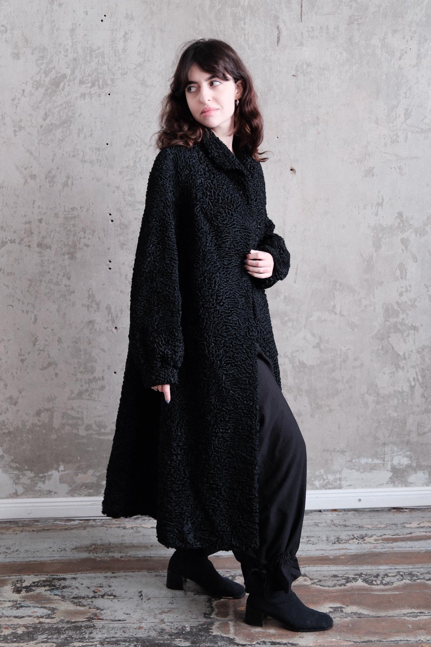 A woman in a black coat stands against a plain gray wall, her gaze directed off to the side.