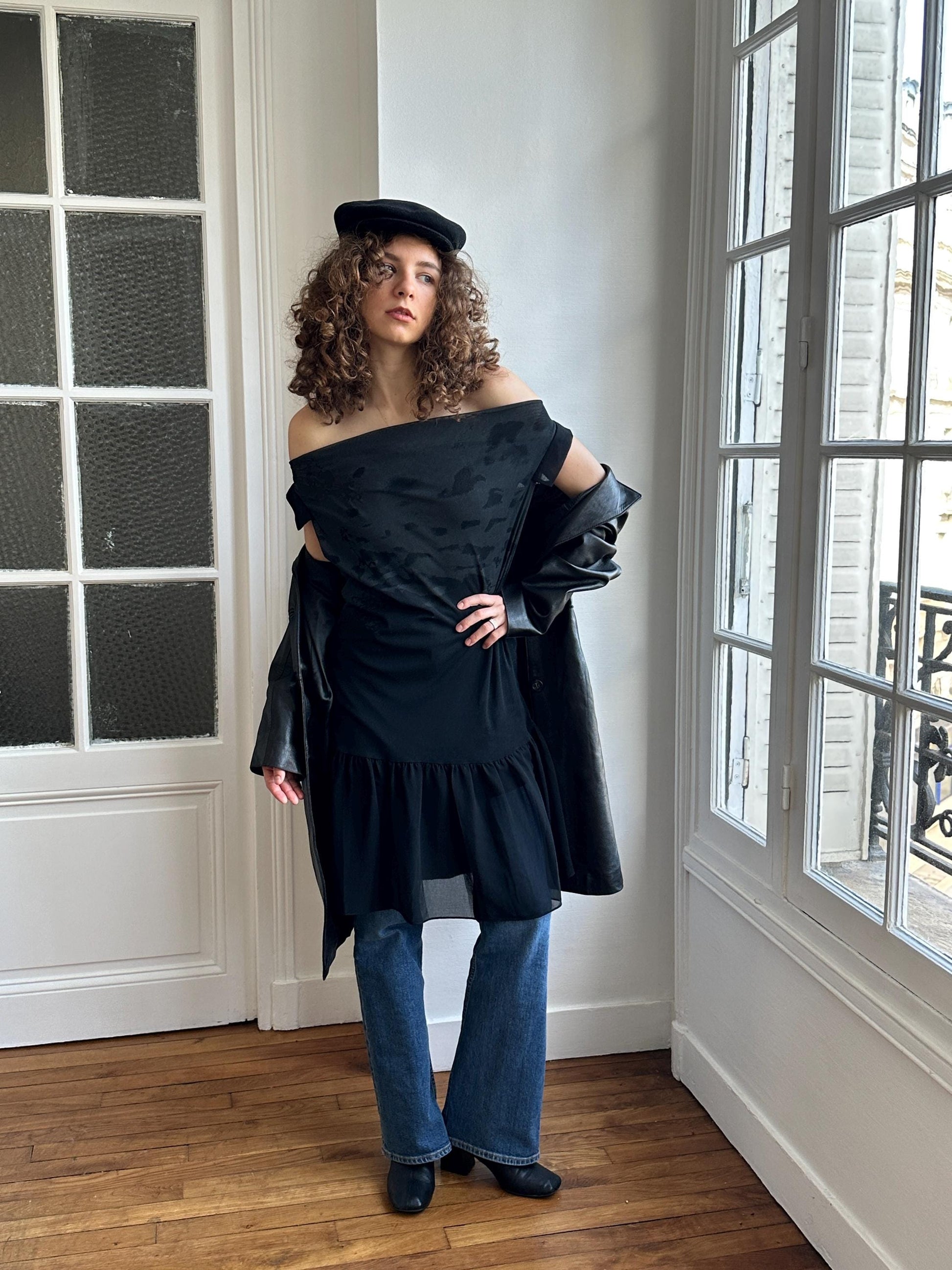 A woman with curly hair wearing a black off-the-shoulder dress and a black hat stands in a room with a window.