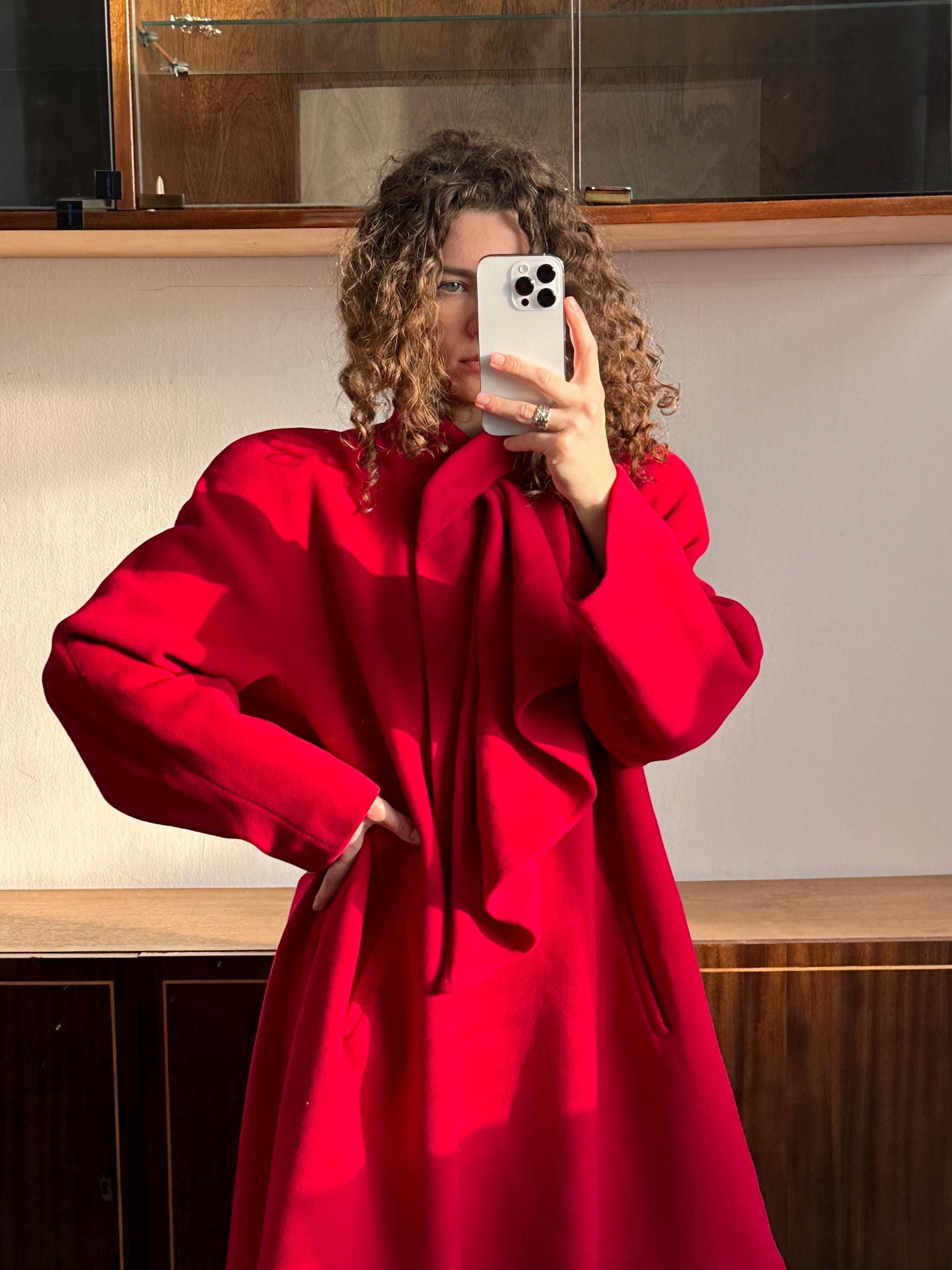 A woman in a red coat is taking a selfie with a white phone, standing in front of a wooden cabinet.
