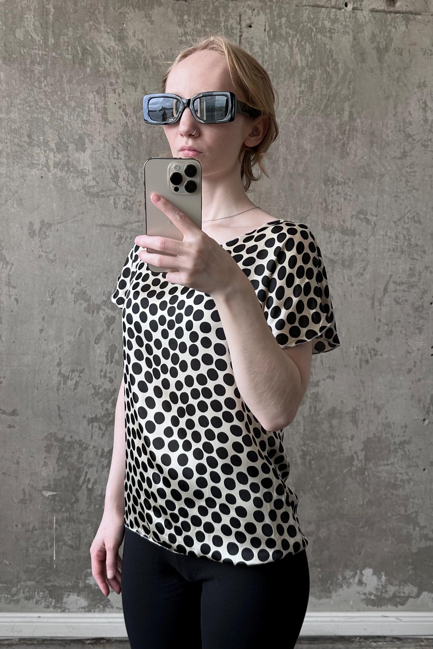 A woman wearing a black and white polka dot blouse and sunglasses is taking a selfie with her iPhone.