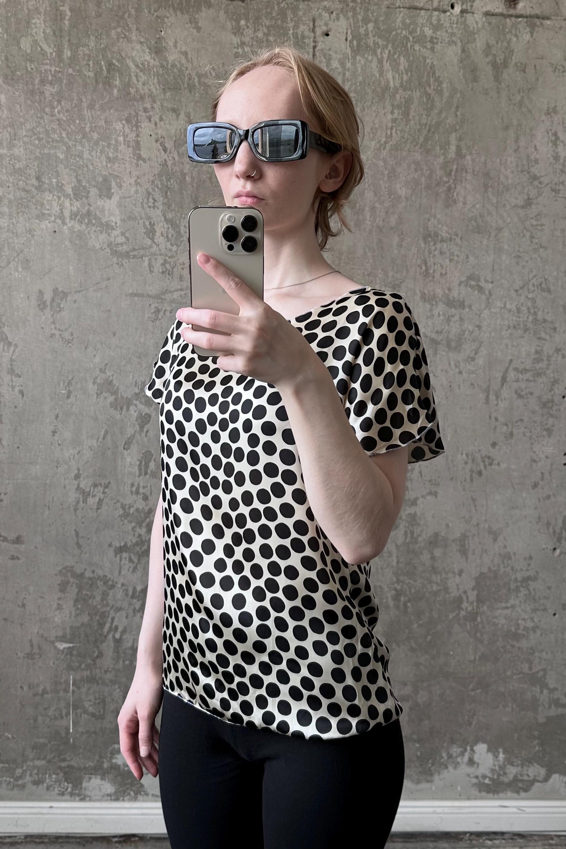A woman wearing a black and white polka dot blouse and sunglasses is taking a selfie with her iPhone.