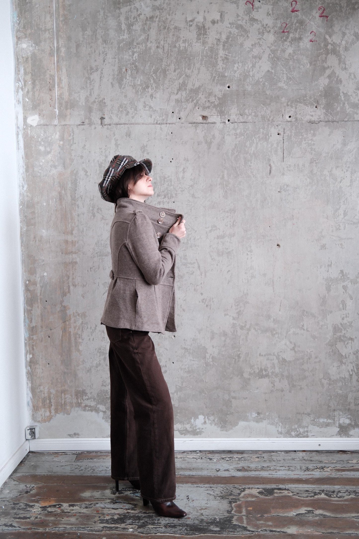A person wearing a brown jacket and hat is standing in a room with a concrete wall and a white baseboard.
