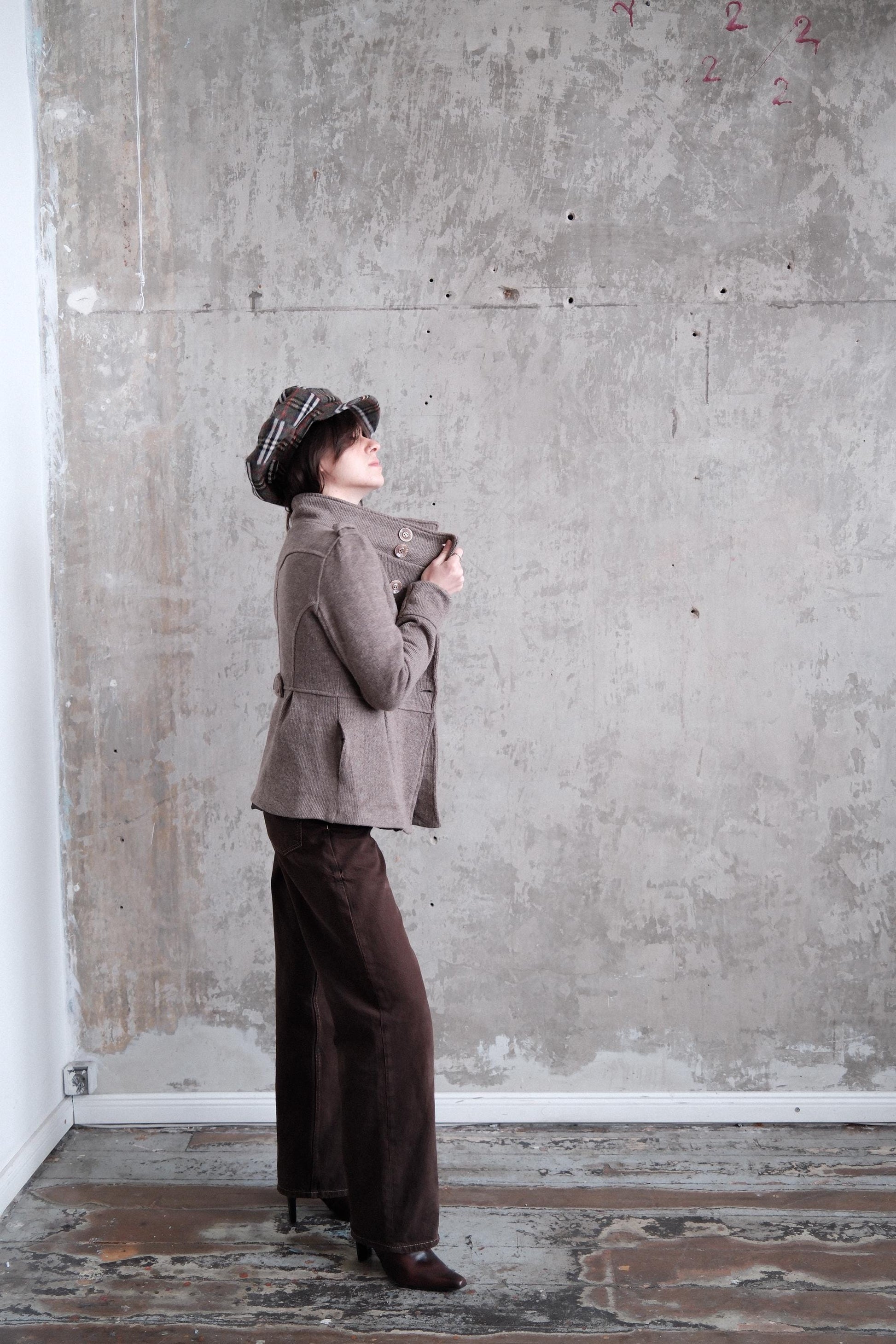 A person wearing a brown jacket and hat is standing in a room with a concrete wall and a white baseboard.