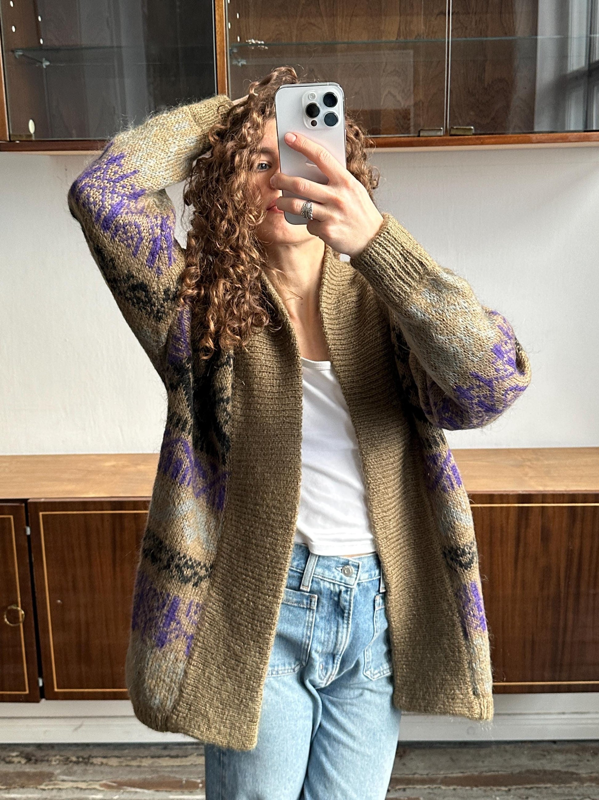 A woman with curly hair is taking a selfie with her iPhone, wearing a brown cardigan with purple and black patterns, and blue jeans.