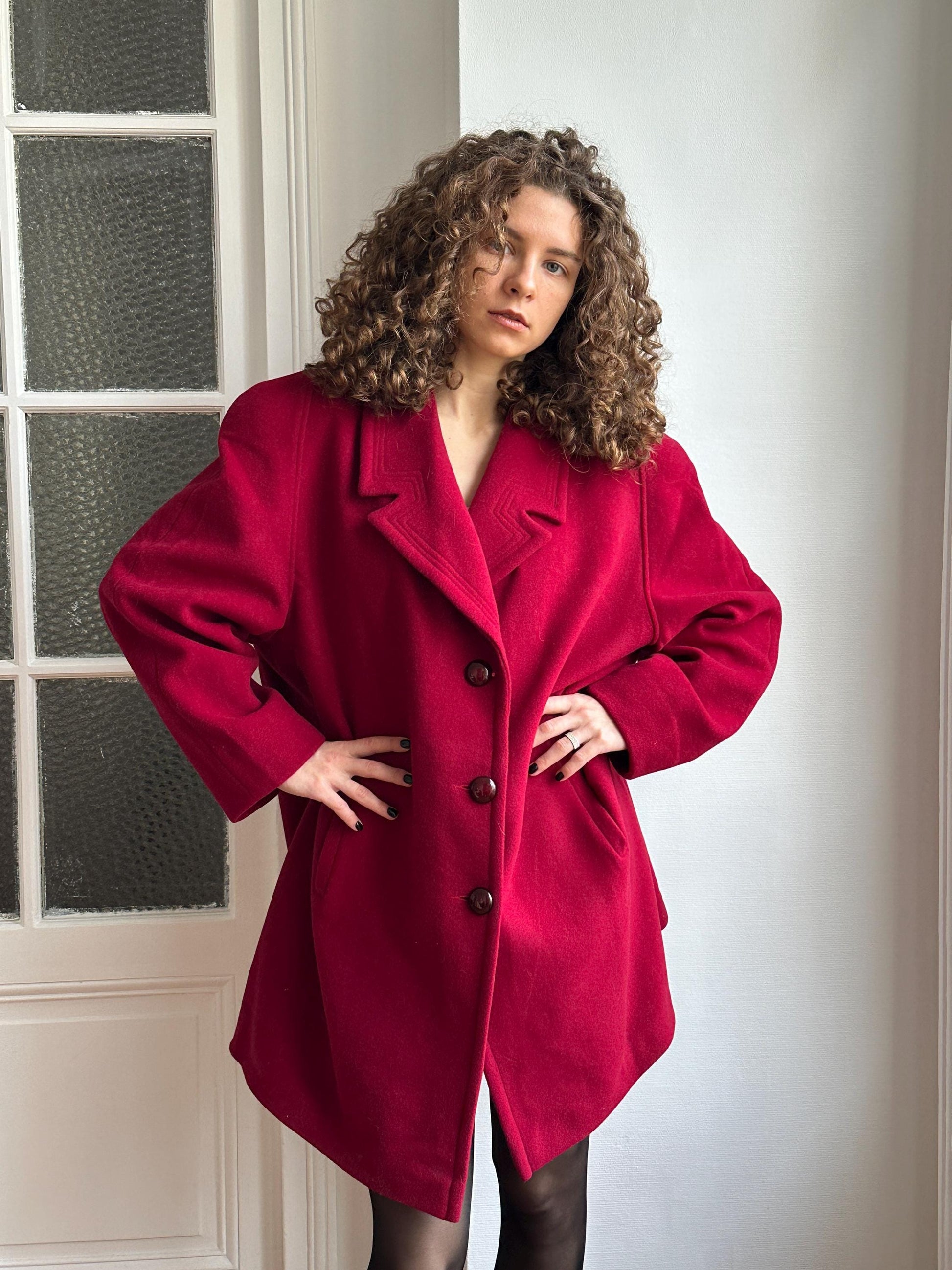 A woman with curly hair wearing a red coat is posing in a relaxed manner.