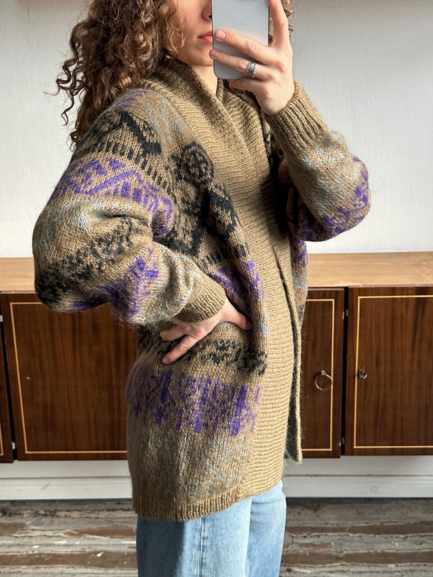 A woman with curly hair is taking a selfie with a phone, wearing a brown and purple patterned cardigan.