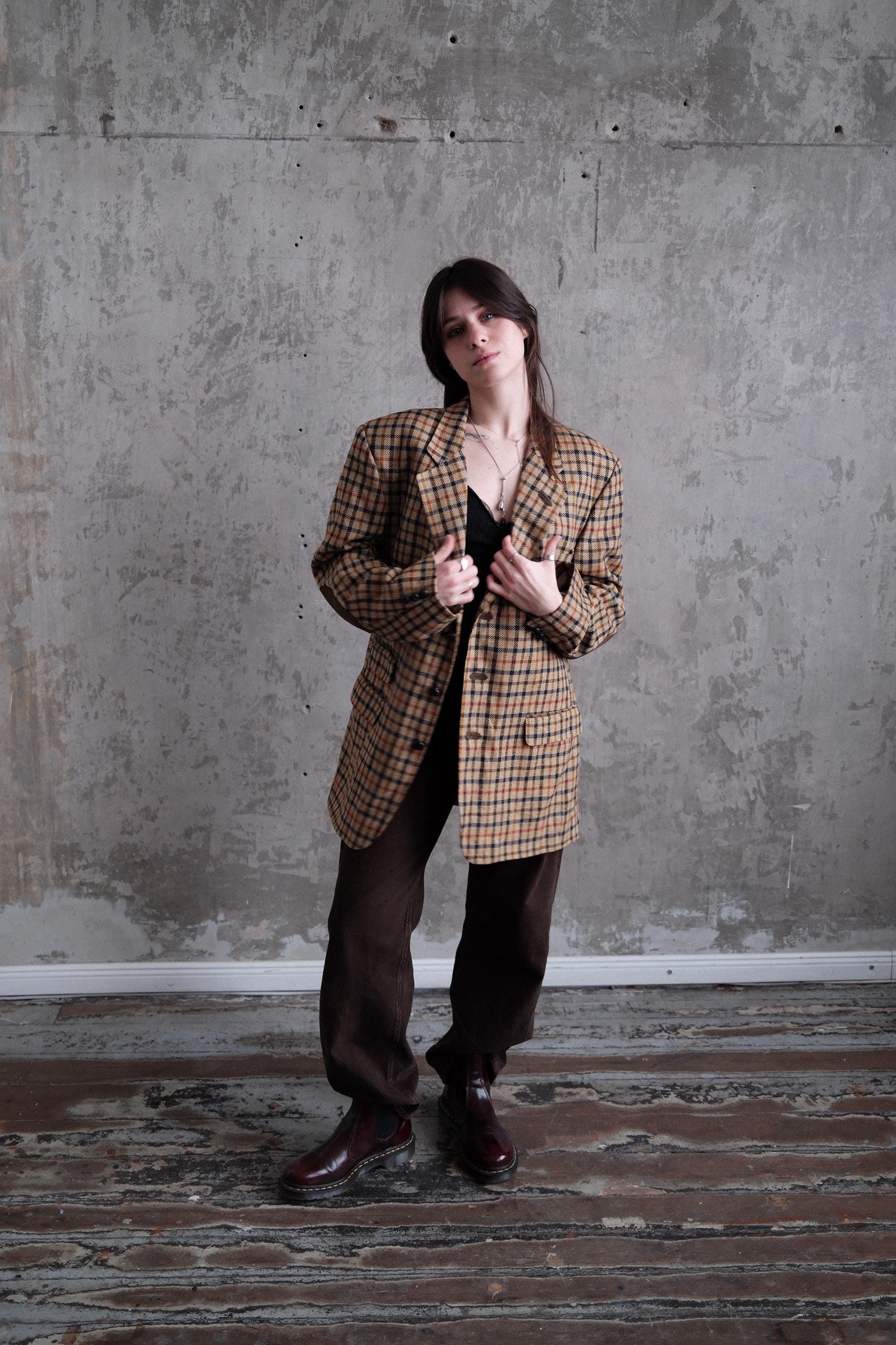 A woman in a plaid jacket stands in a room with a concrete wall and wooden floor.
