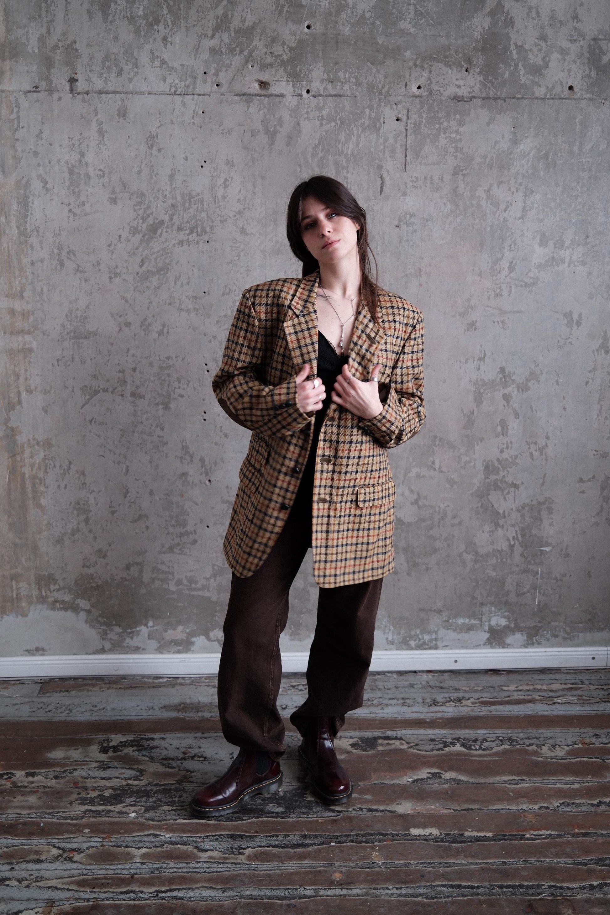 A woman in a plaid jacket stands in a room with a concrete wall and wooden floor.