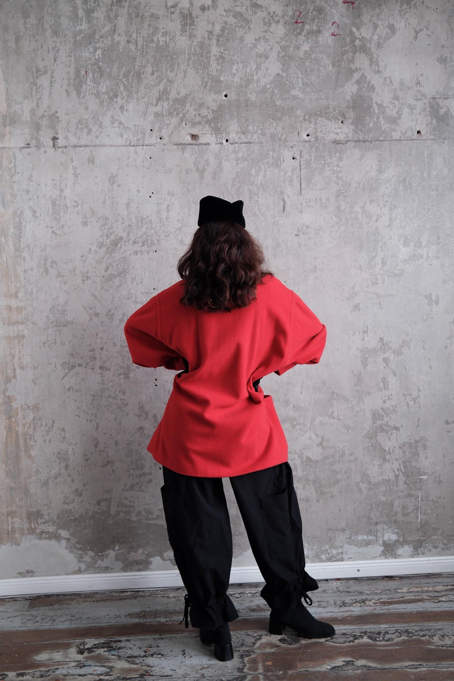 A person wearing a red jacket and black pants is standing in front of a concrete wall.