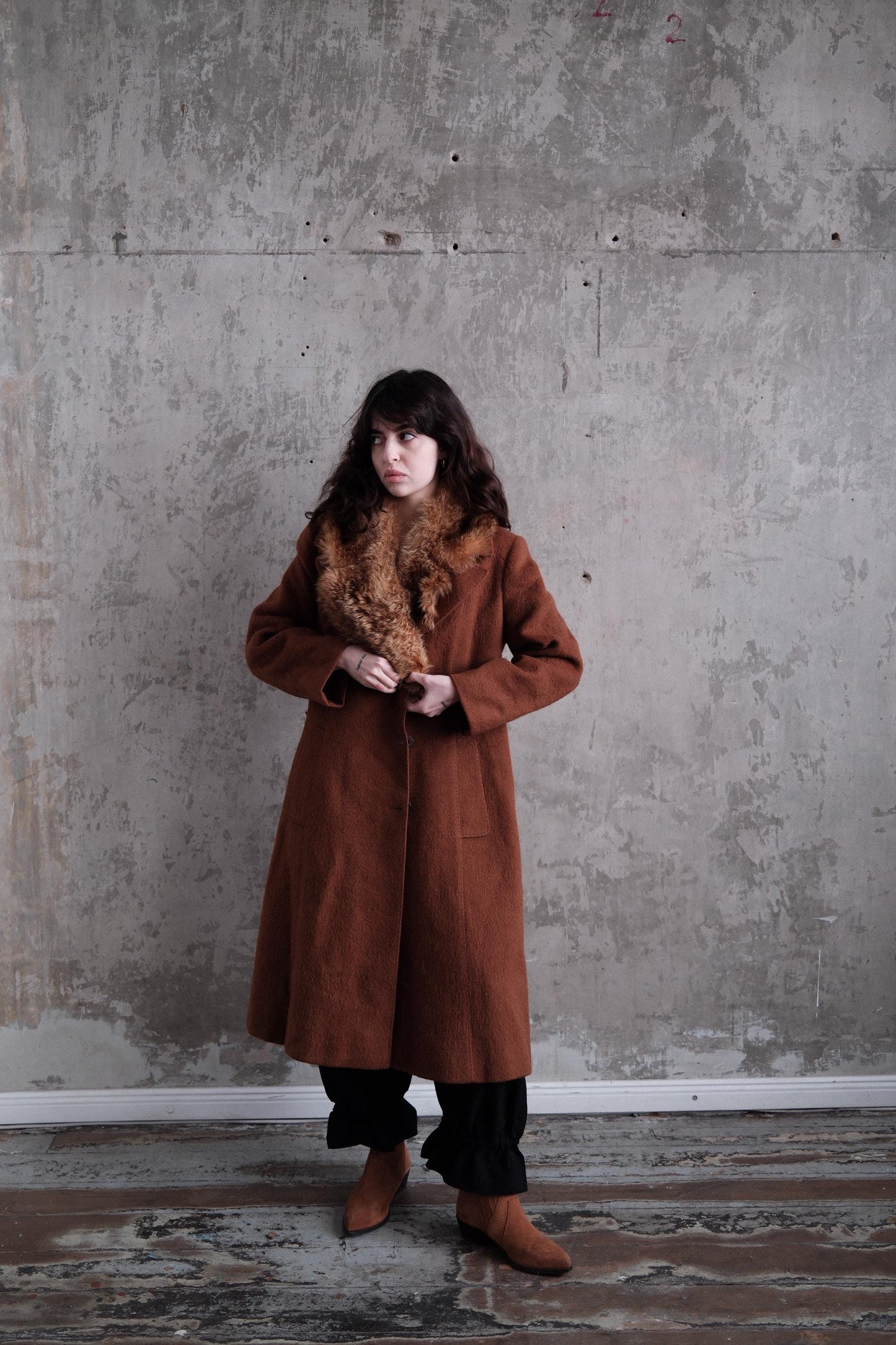 A woman in a brown coat stands against a concrete wall, her gaze directed off to the side.