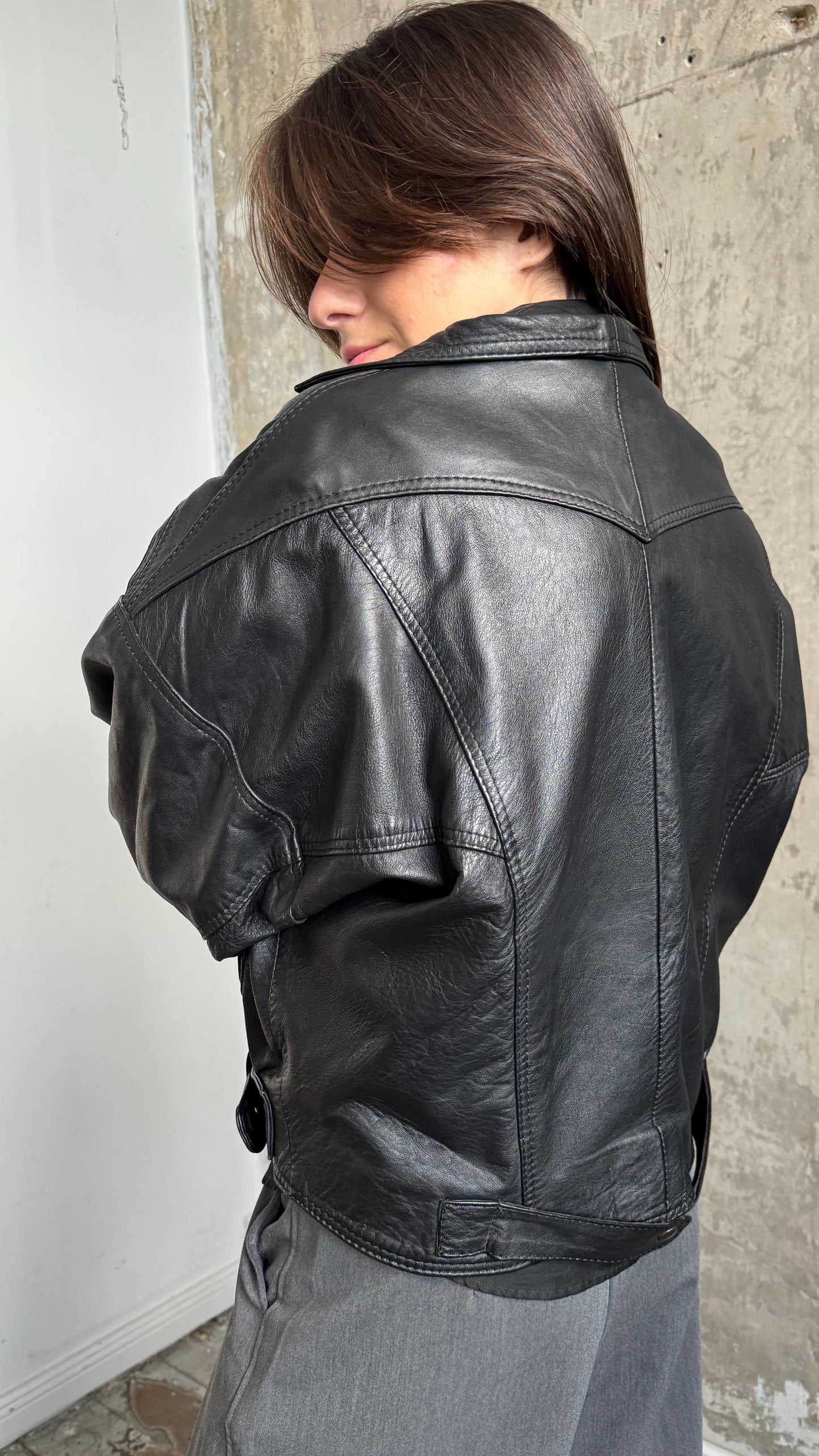 A woman wearing a black leather jacket stands in front of a concrete wall.