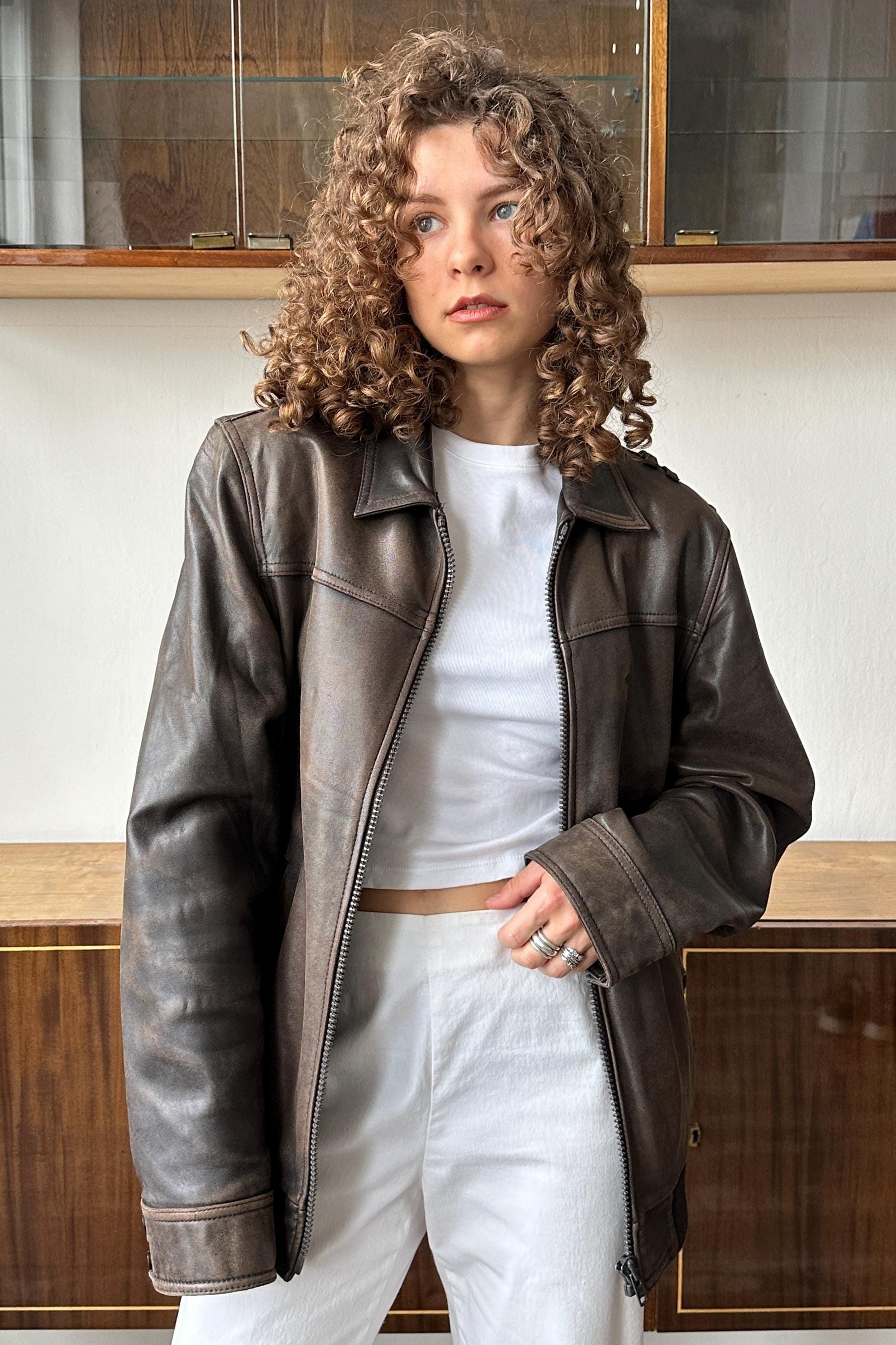 A woman with curly hair wearing a brown leather jacket and white pants stands in front of a wooden cabinet.