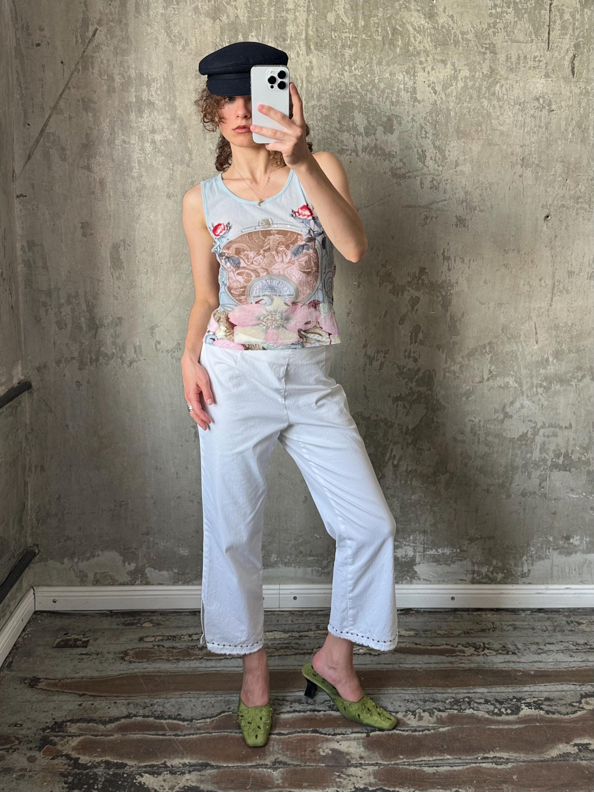A woman is taking a selfie with a smartphone, wearing a floral tank top, white pants, and green shoes.