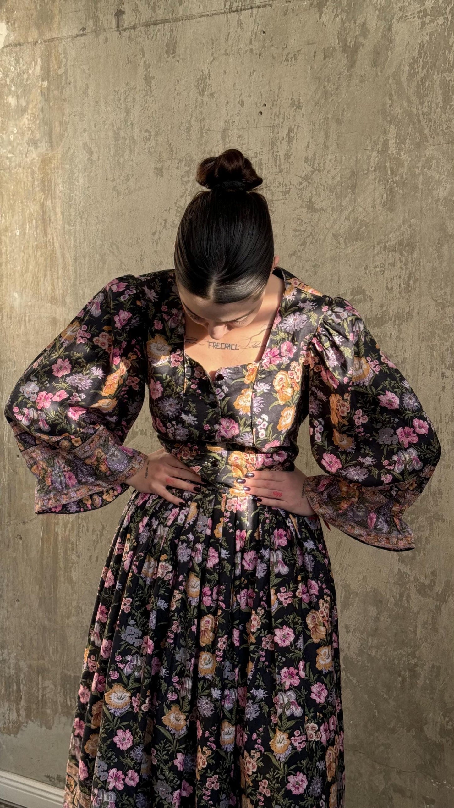 A woman wearing a floral print dress stands in front of a concrete wall, her hands on her hips.