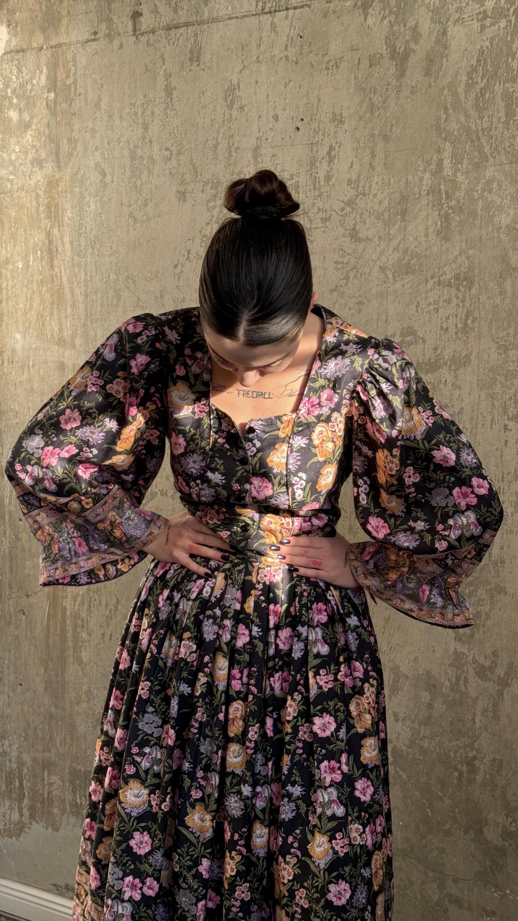A woman wearing a floral print dress stands in front of a concrete wall, her hands on her hips.