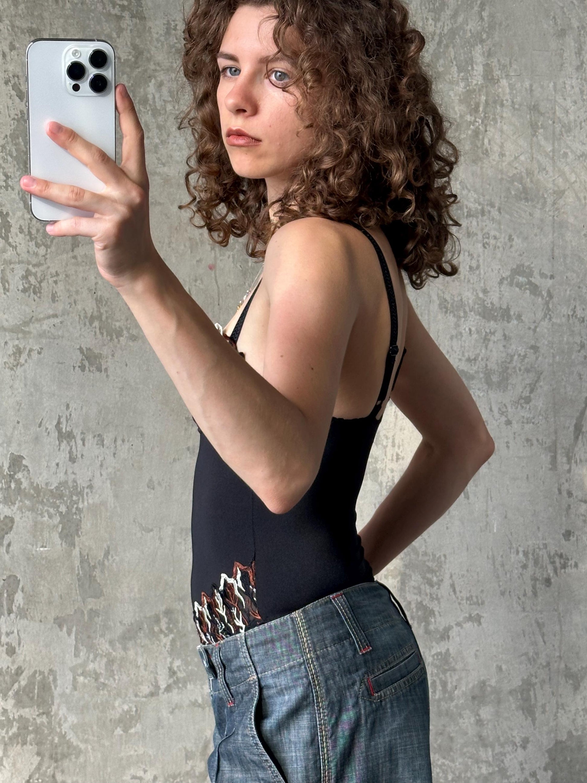A woman with curly hair is taking a selfie with a smartphone, wearing a black tank top and blue jeans.
