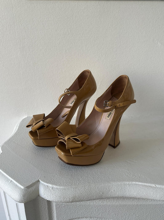 A pair of tan high-heeled shoes with bows on the toe and heel, placed on a white surface.