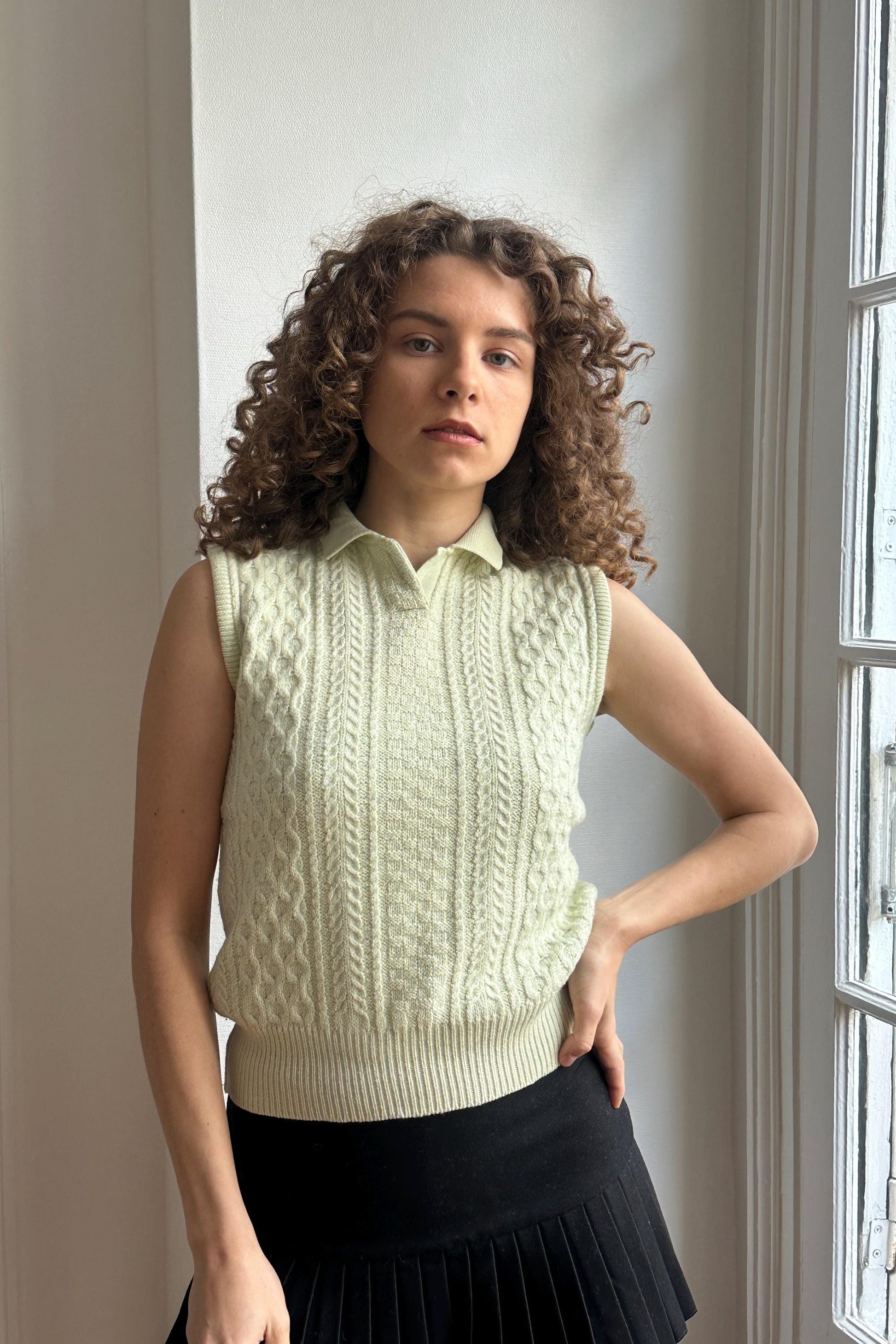 A young woman with curly hair stands in front of a window, wearing a white sweater vest and a black skirt.