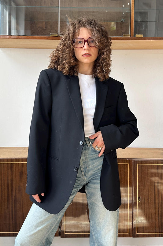 A woman with curly hair wearing a black blazer, white shirt, and jeans stands in front of a wooden cabinet.