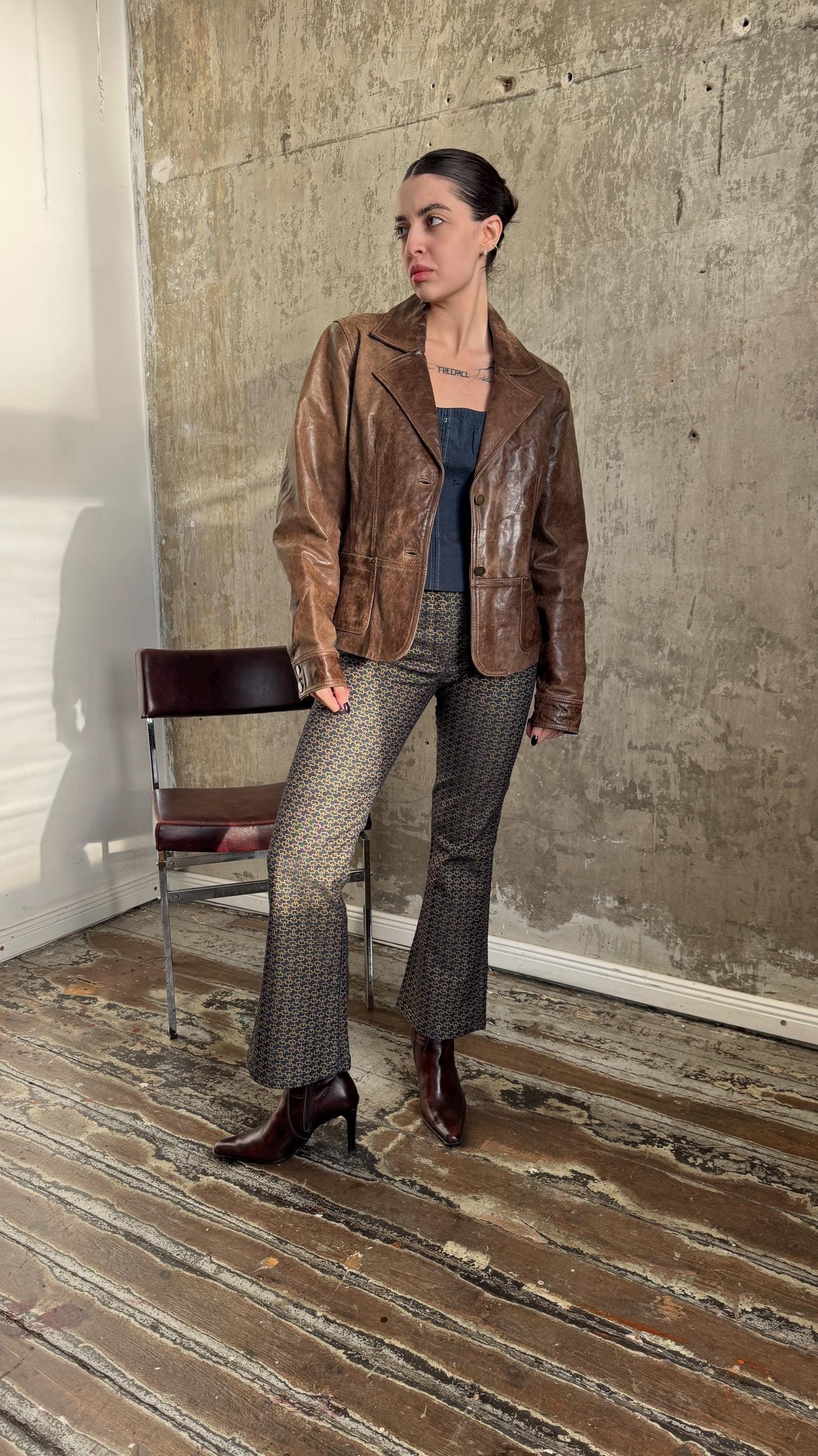 A woman wearing a brown leather jacket and patterned pants stands in a room with a wooden floor and a chair.