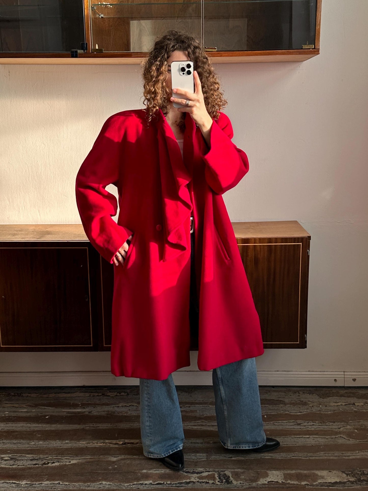 A person wearing a red coat is taking a selfie with a smartphone in front of a wooden cabinet.