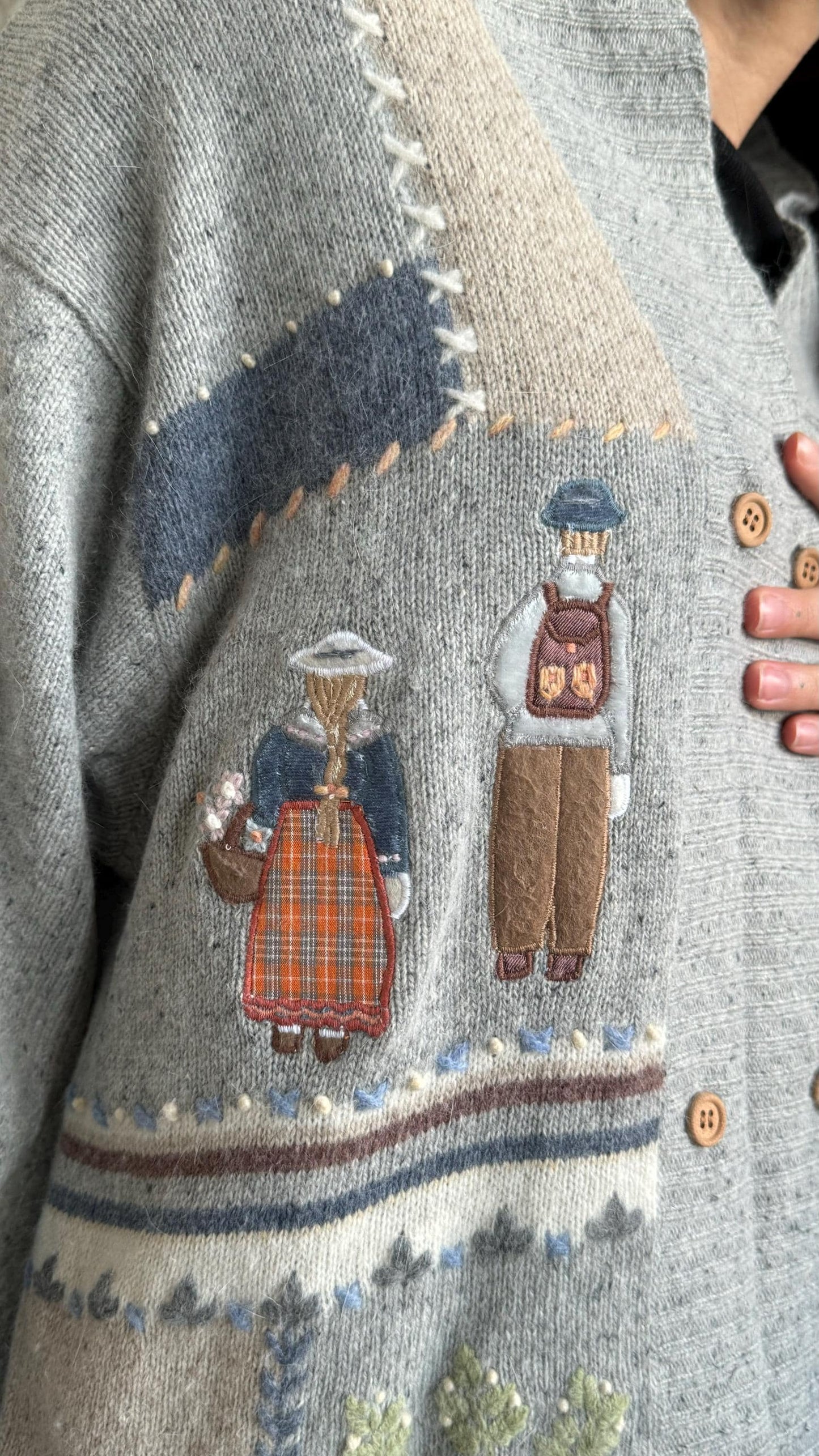 a close-up of a gray sweater with a patchwork design featuring two figures, one wearing a plaid skirt and the other wearing a hat.