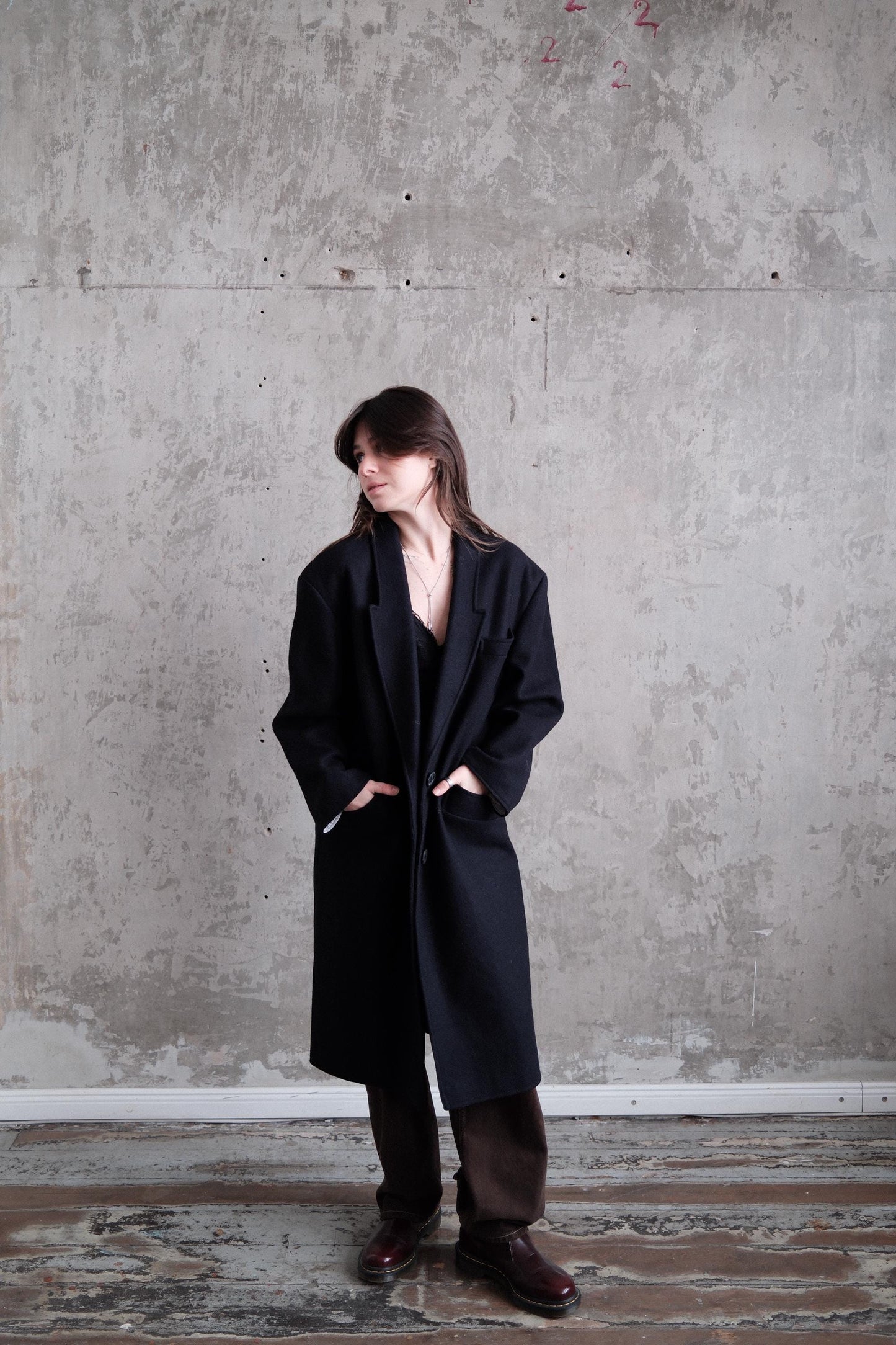 A person wearing a black coat stands in a room with a concrete wall.
