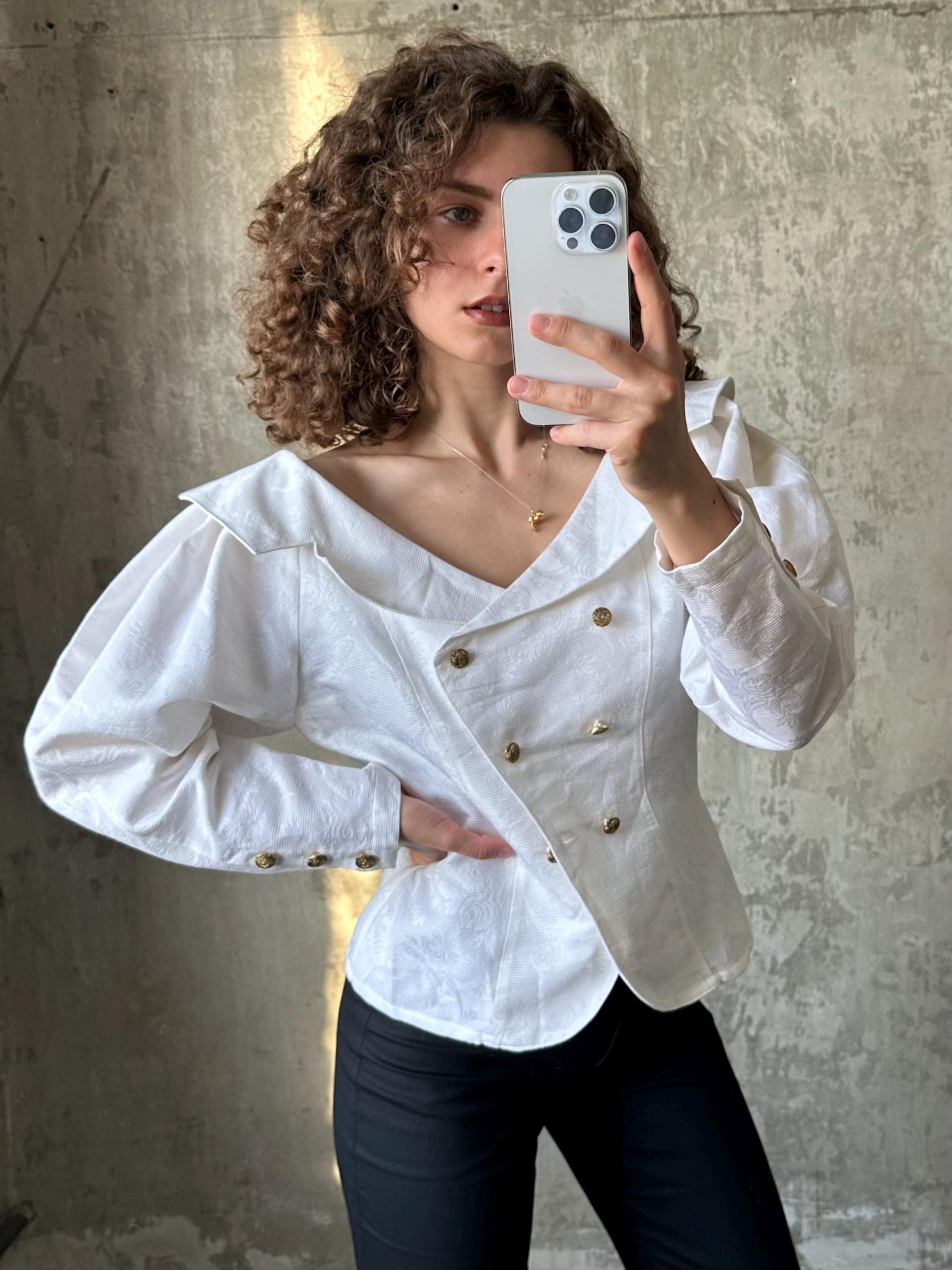 A woman with curly hair is taking a selfie with a white smartphone, wearing a white blazer with gold buttons.