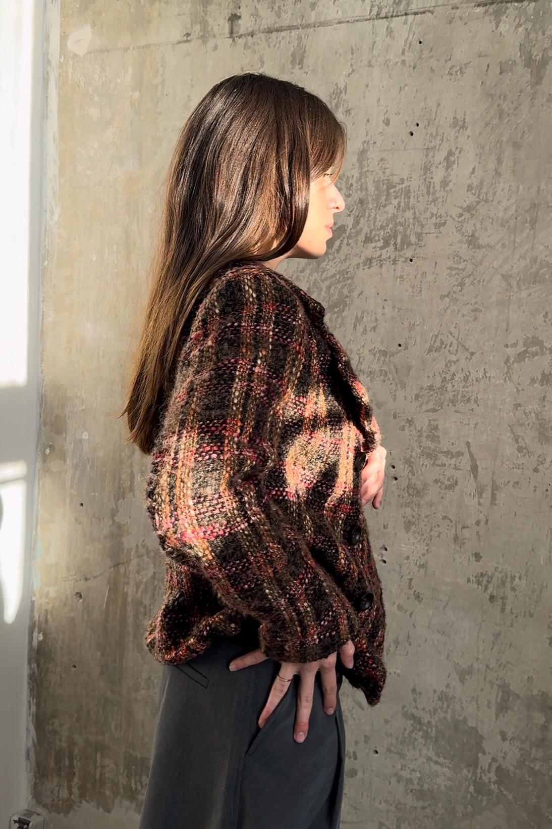A woman with long brown hair stands in front of a concrete wall, wearing a brown and red plaid sweater.