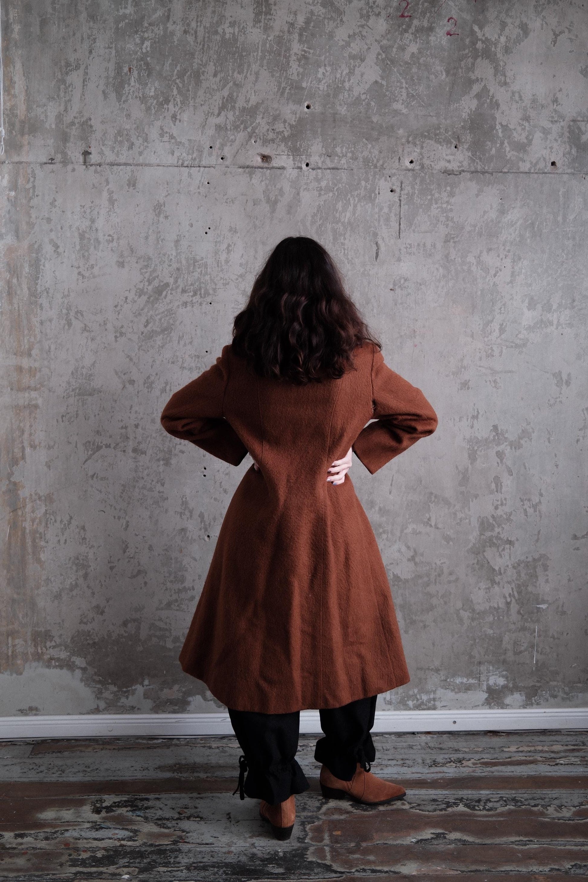 A person wearing a brown coat stands in front of a concrete wall, with their back to the camera.