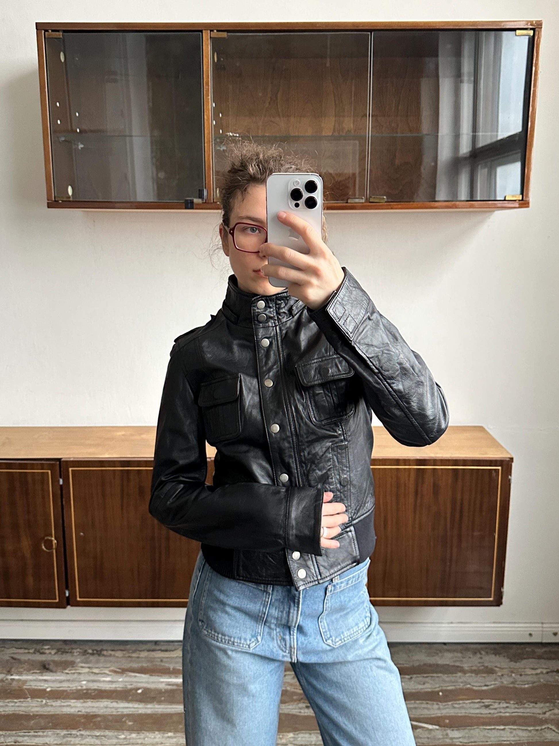 A person is taking a selfie with a smartphone, wearing a black leather jacket and blue jeans, in front of a wooden cabinet.