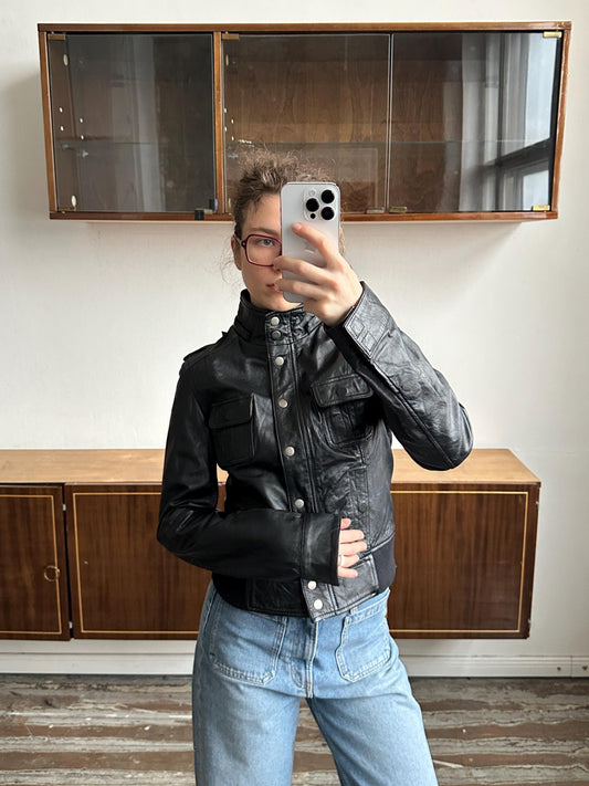 A person is taking a selfie with a smartphone, wearing a black leather jacket and blue jeans, in front of a wooden cabinet.