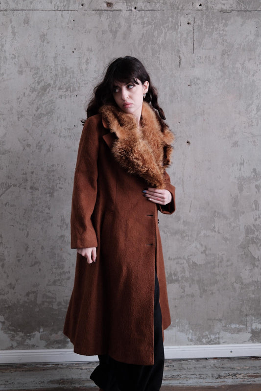 A woman wearing a brown coat with fur trim is standing against a concrete wall.