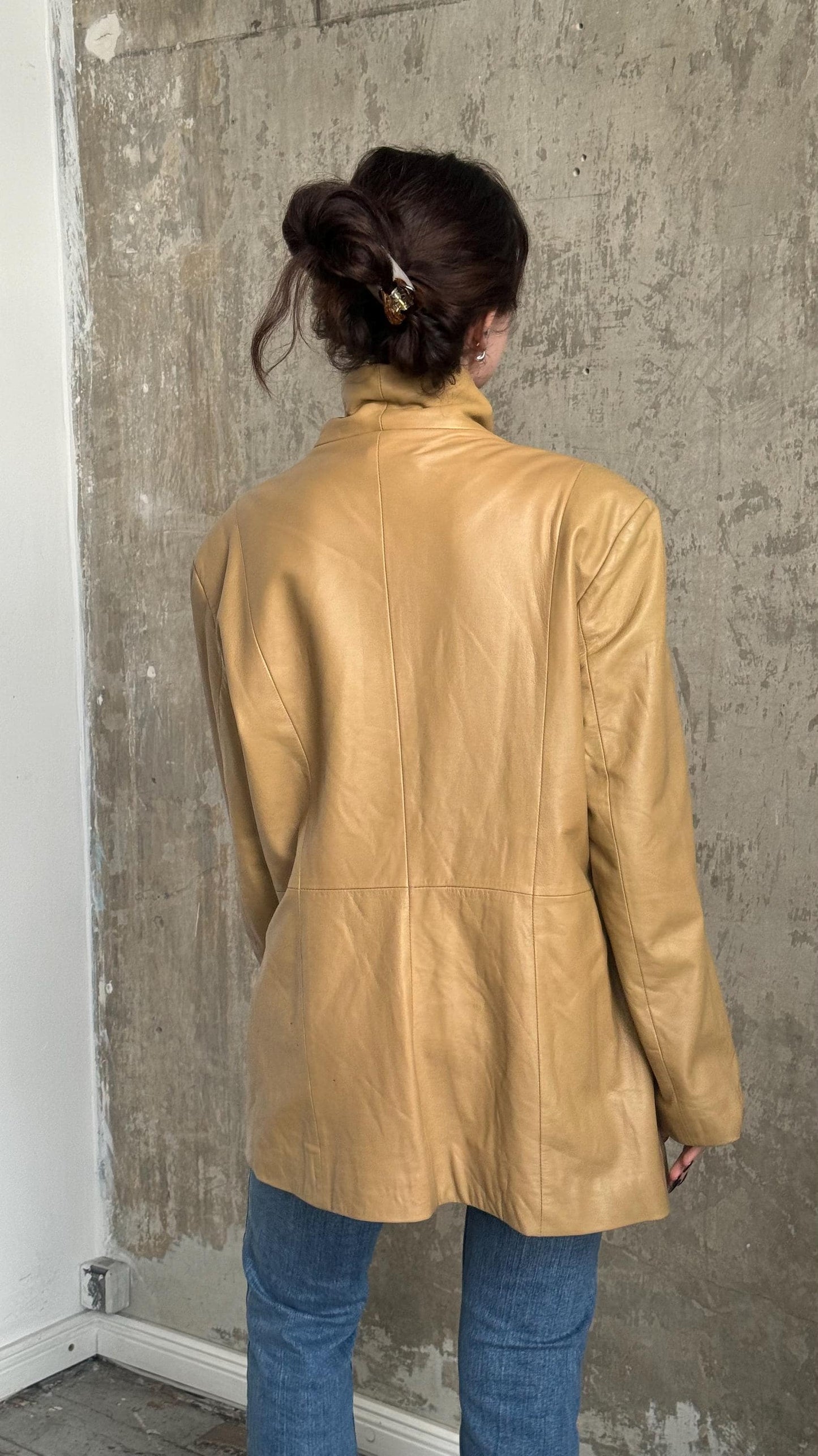 a person wearing a tan leather jacket and blue jeans, standing against a concrete wall.