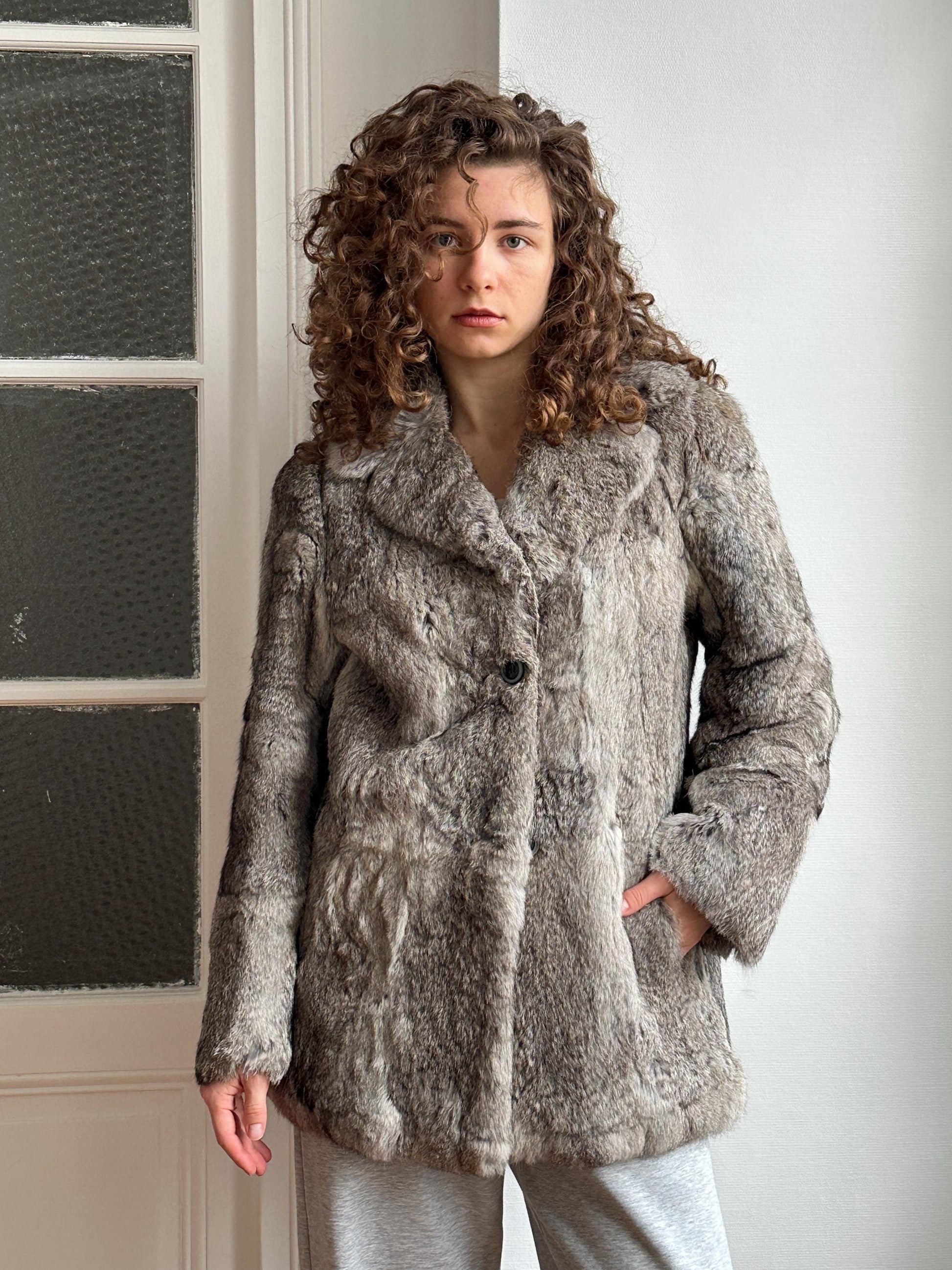 A young woman with curly hair stands in front of a white door, wearing a gray fur coat.