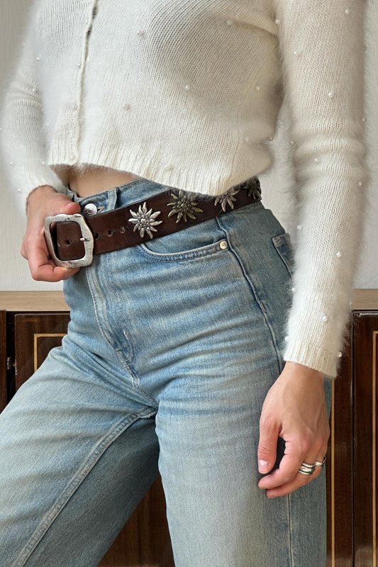 A person wearing a white sweater, blue jeans, and a brown belt with metal studs stands in front of a wooden cabinet.