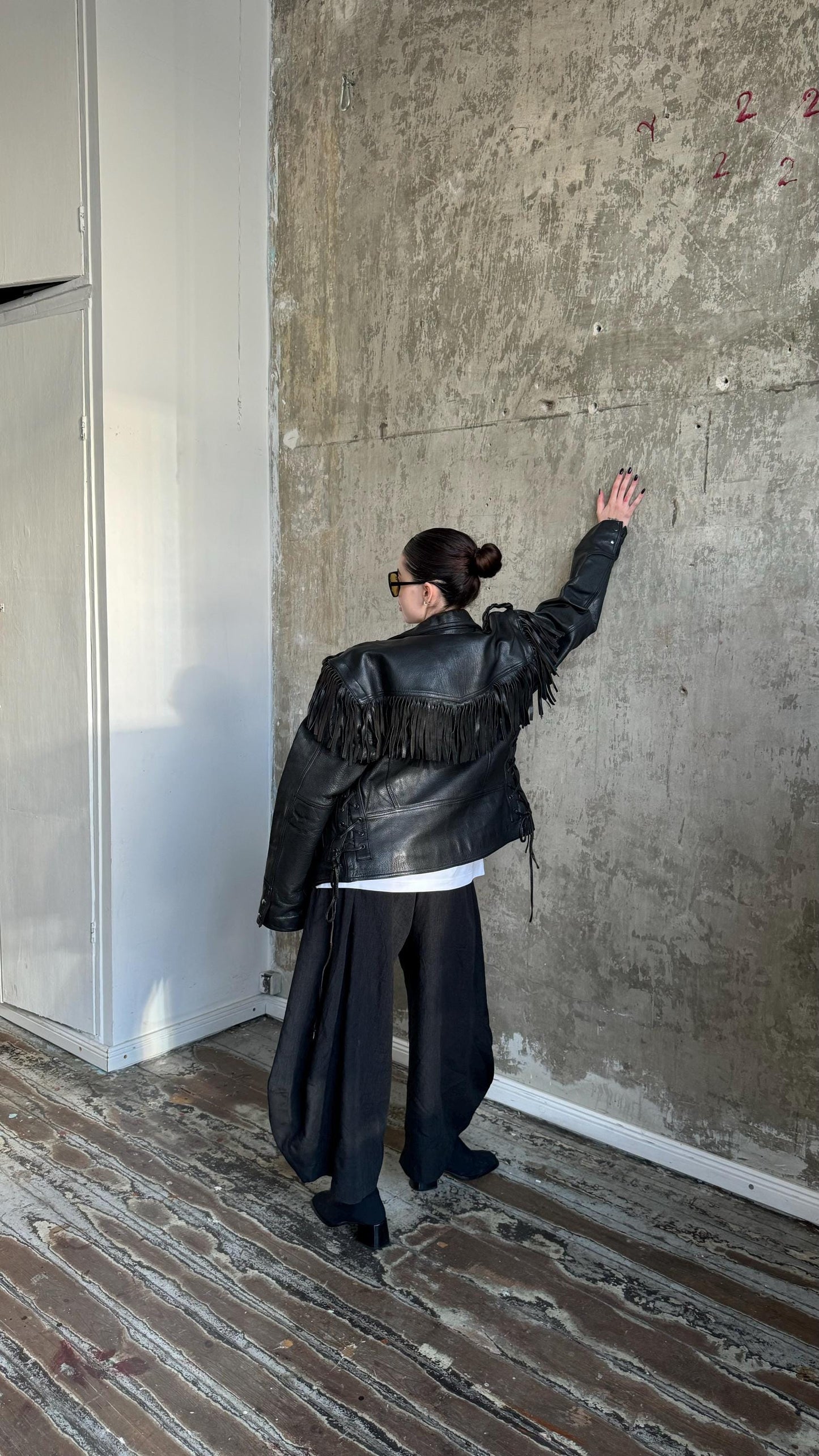 A person wearing a black leather jacket with fringe and sunglasses stands in a room with a concrete wall and wooden floor.