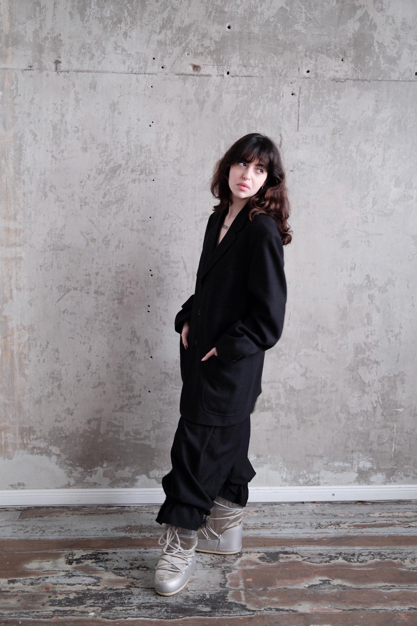 A woman in a black outfit stands against a concrete wall, her gaze directed downwards.