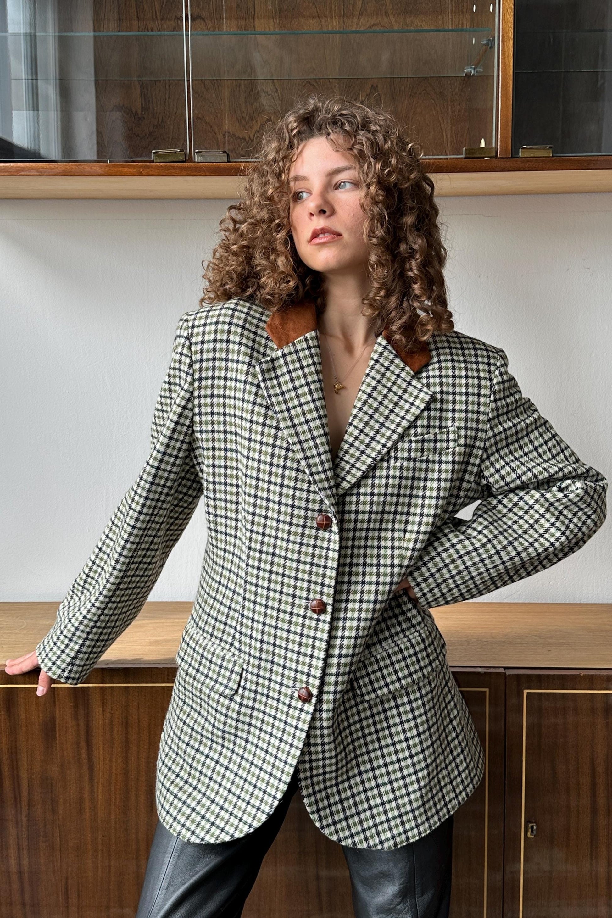 A woman with curly hair wearing a checkered jacket stands in front of a wooden cabinet.
