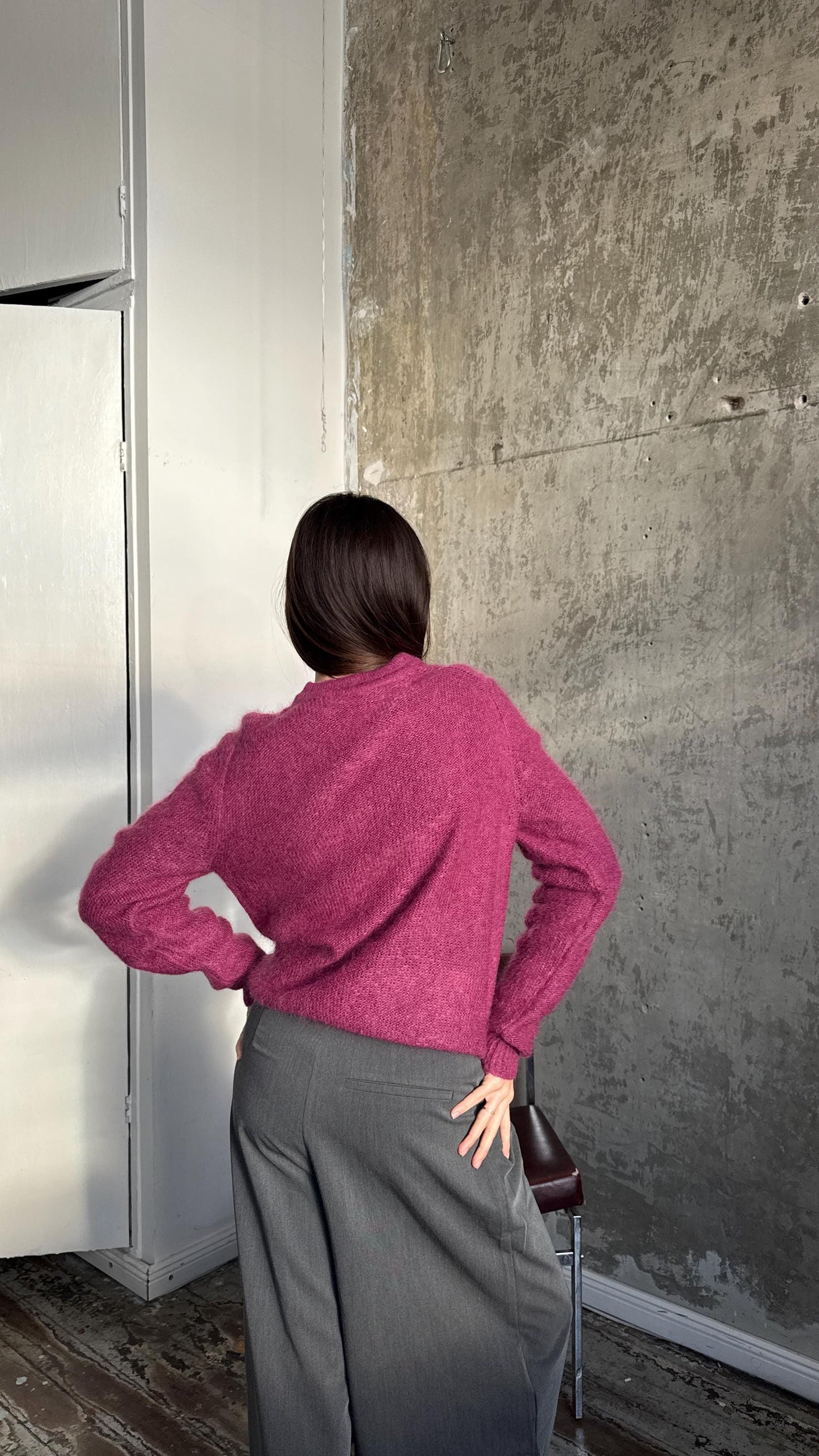 A person wearing a pink sweater stands in front of a concrete wall, with their back to the camera.