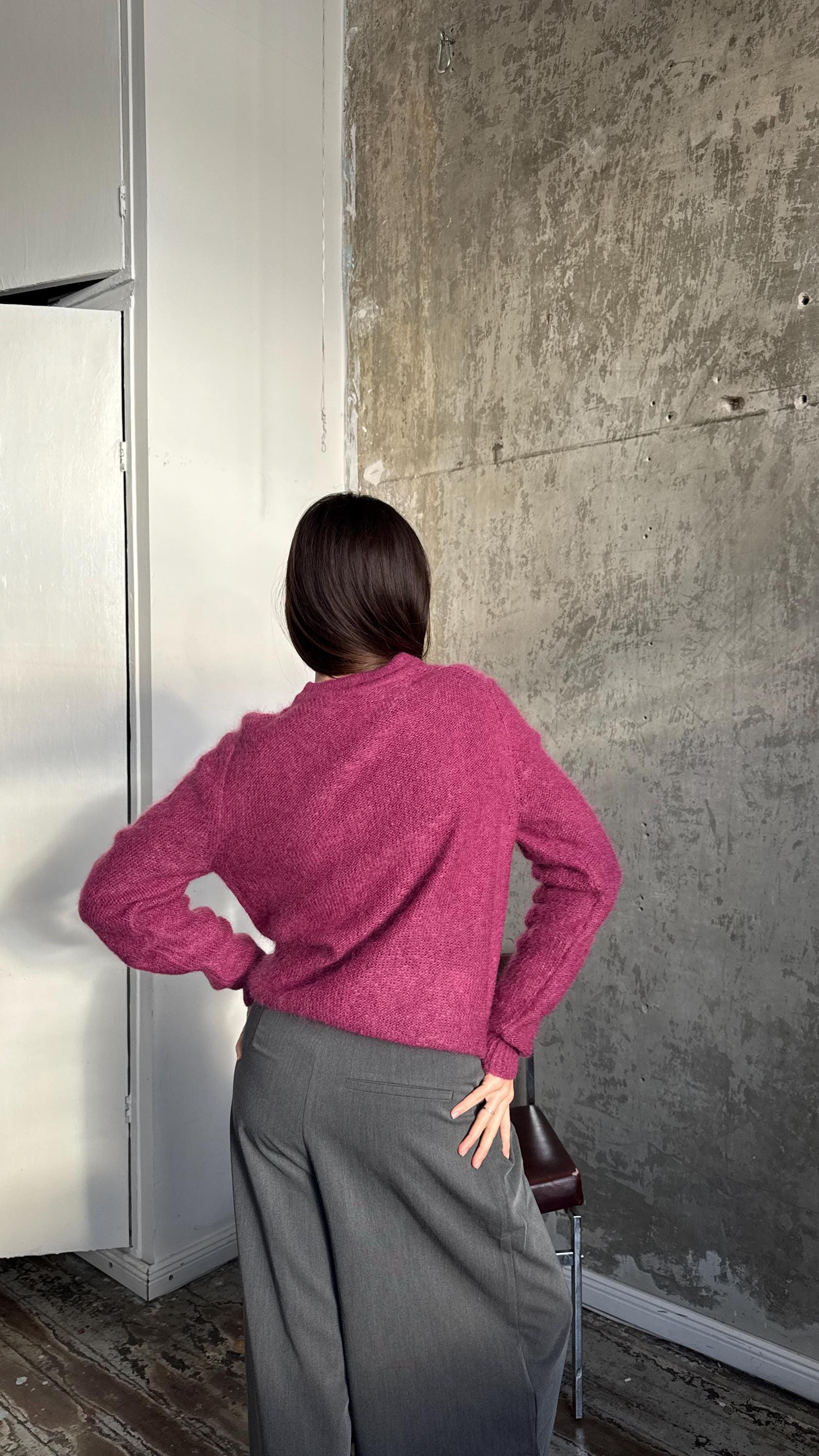 A person wearing a pink sweater stands in front of a concrete wall, with their back to the camera.