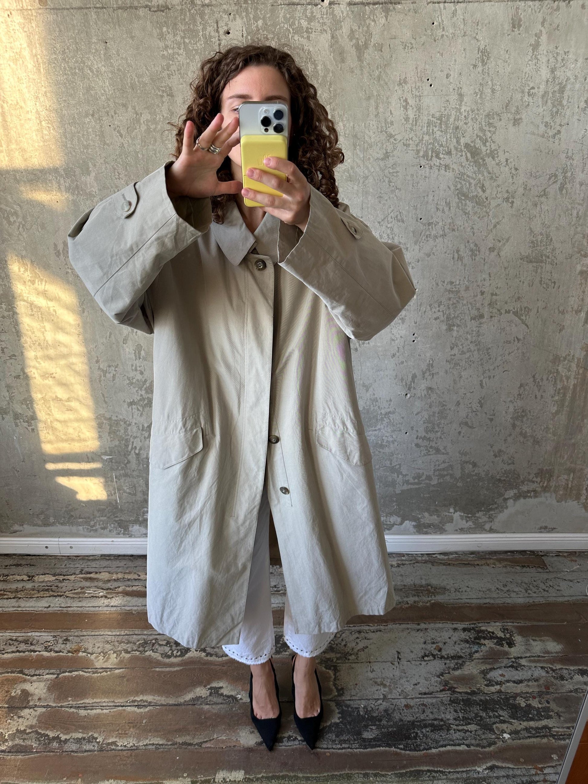 A woman in a beige trench coat is taking a selfie with her phone, standing in front of a concrete wall.