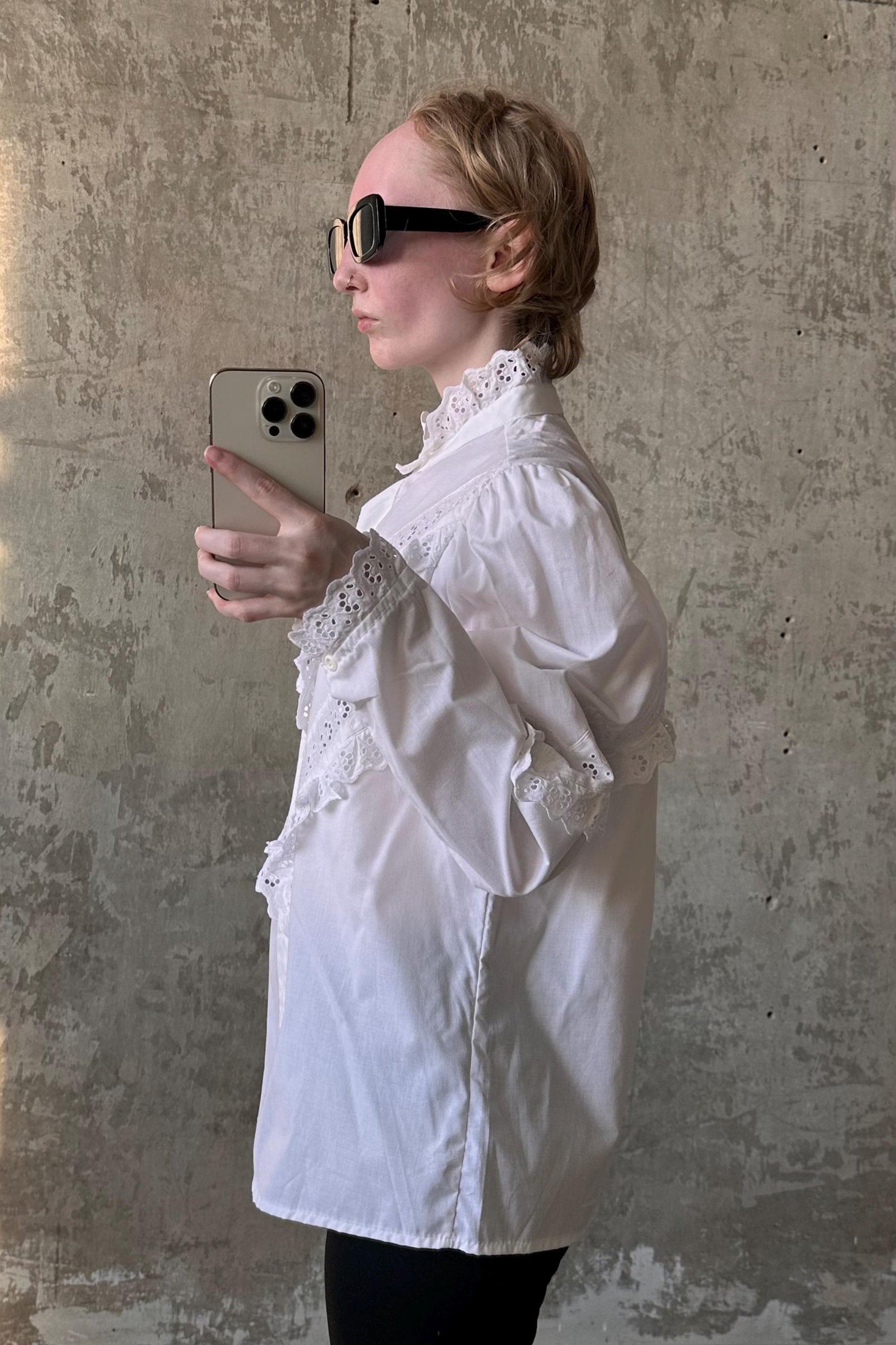 A woman wearing a white blouse and sunglasses is taking a selfie with a smartphone.