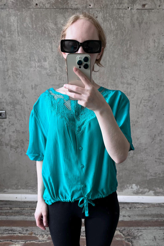 A woman wearing a turquoise blouse and sunglasses is taking a selfie with her iPhone.