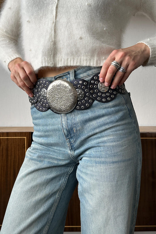 A person wearing a white sweater and blue jeans, with their hands on their hips, and a large, decorative belt with a metal buckle.