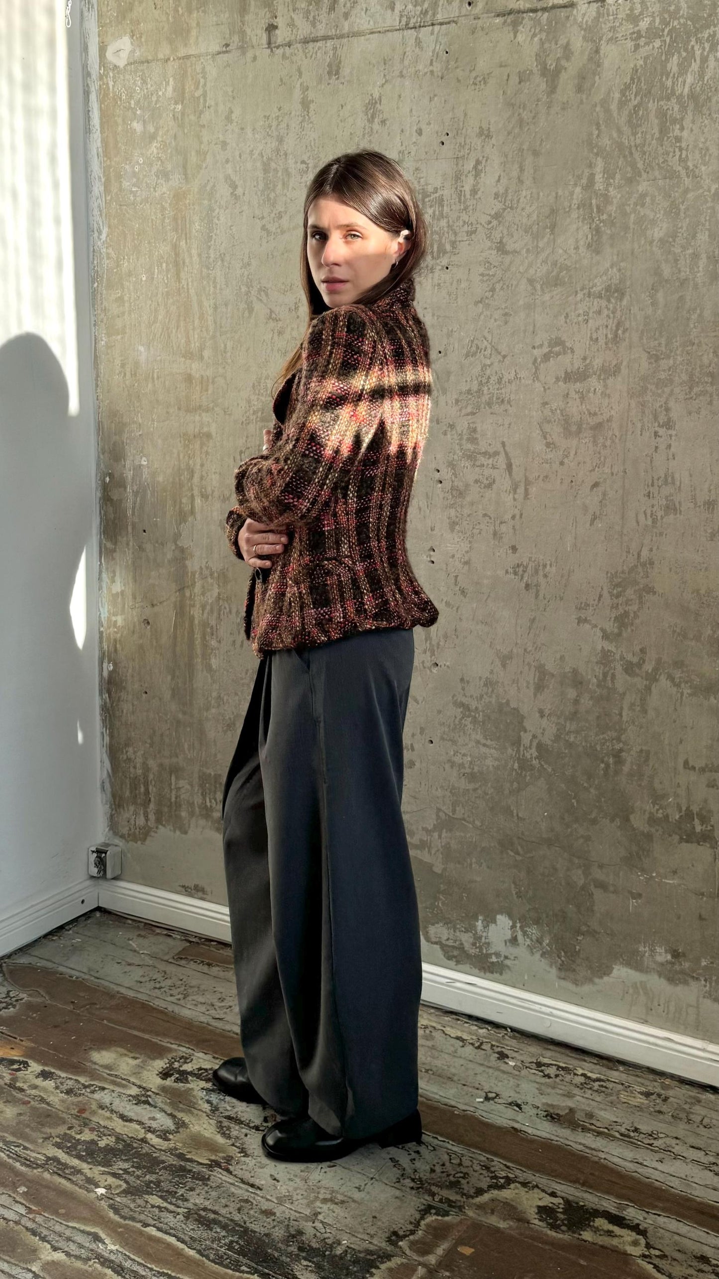 A woman wearing a brown and black patterned jacket stands in a room with a concrete wall.