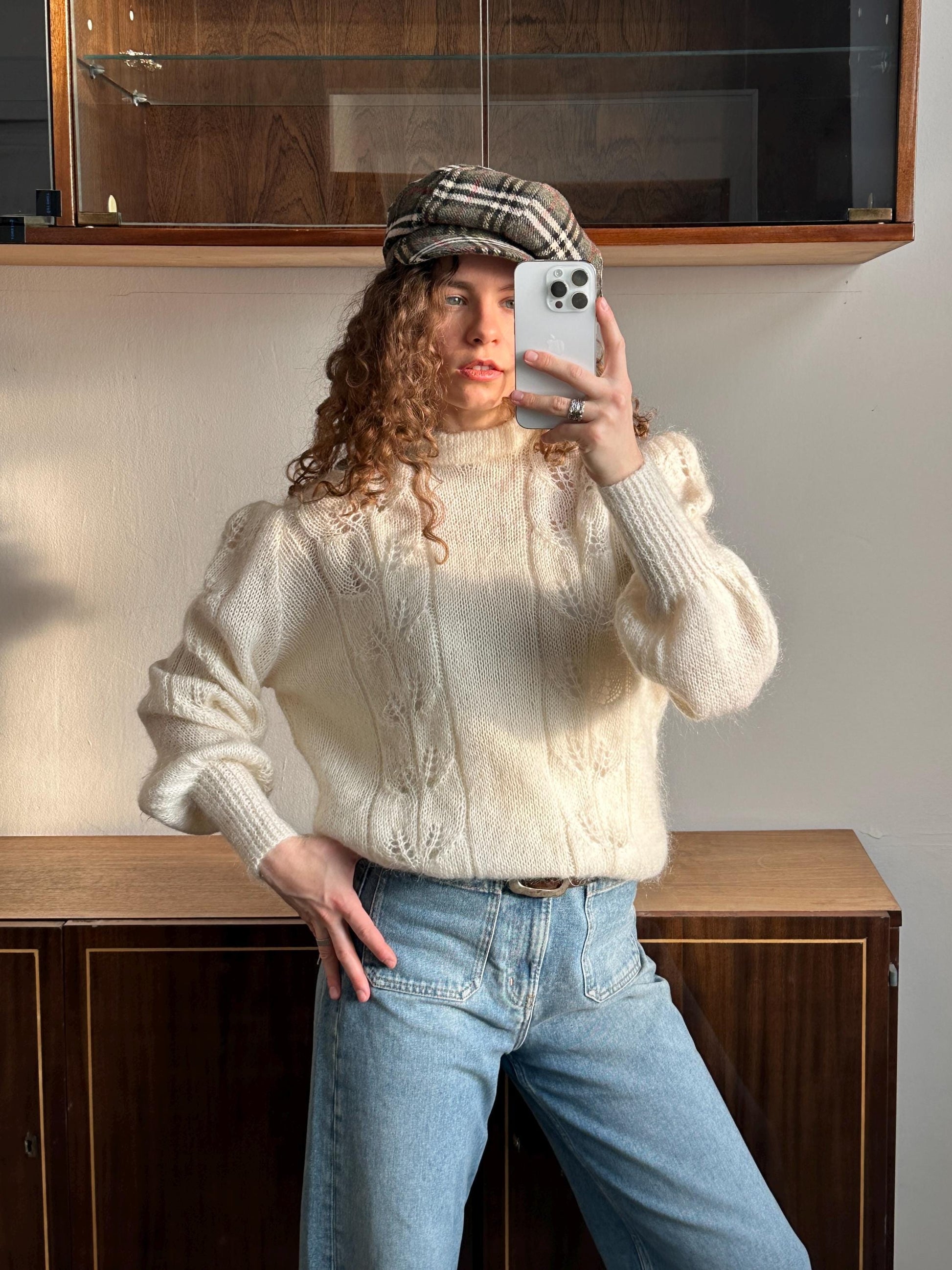 A woman with curly hair is taking a selfie with a white iPhone, wearing a white sweater and blue jeans.