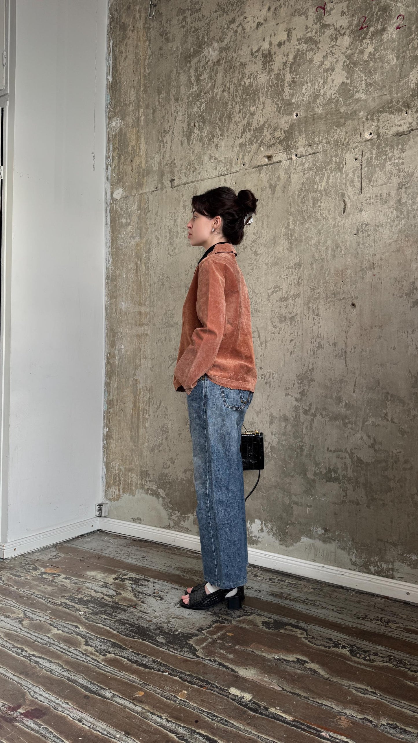 A woman in a brown jacket and blue jeans stands in a room with a concrete wall and wooden floor.