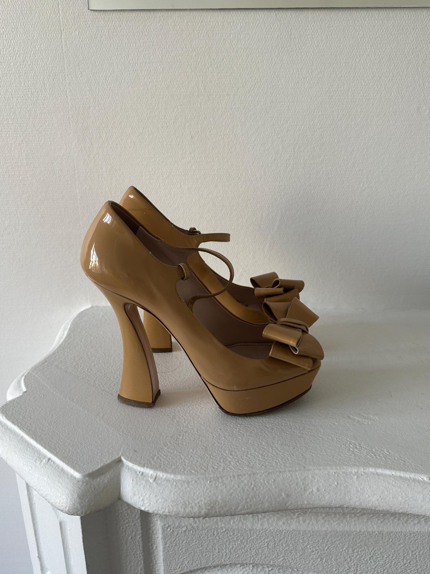 A pair of tan high-heeled shoes with bows on the toe are displayed against a white wall.